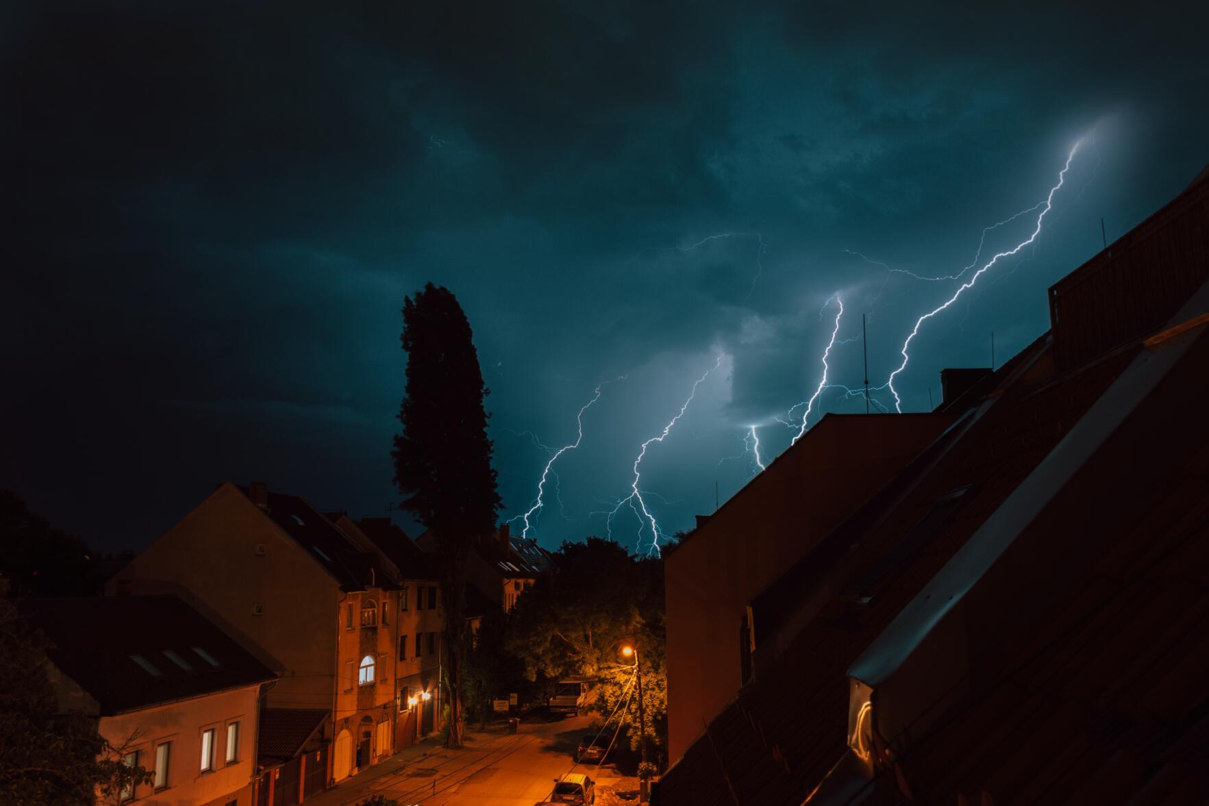 Preparing Your Roof Before a Storm. Image of bolts of light in the nighttime sky, above several buildings.