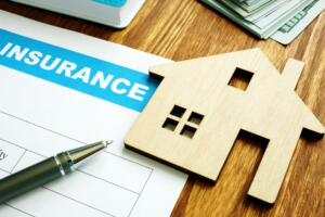 What Does Homeowners Insurance Cover. Image of a wooden home cutout sitting on top of insurance paperwork. Money and an ink pen sit around the house cutout.